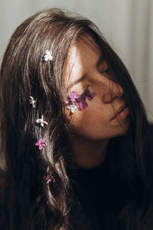 Beautiful sensual woman with lilac petals on face and hair in sunlight. Moody portrait of young brunette with flowers on face in light and shadow. Skin care and natural cosmetics conceptの写真素材