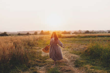 Stylish woman with straw hat dancing at oat field in sunset light. Atmospheric happy moment. Young female in rustic linen dress relaxing in evening summer countryside, rural lifeの写真素材