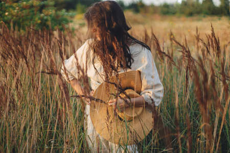 Stylish boho woman with straw hat posing among wild grasses in evening. Summer delight and travel....