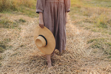 Woman with hat in hand walking barefoot on straw field in sunset light, close up. Rural slow life....