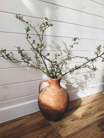 Blooming cherry branch in rustic vase on sunny wooden background in scandinavian room. Home Decor and spring details. Vertical phone photoの写真素材