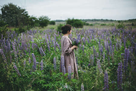 Stylish woman in rustic dress gathering lupine bouquet in meadow, atmospheric moment. Cottagecore aesthetics. Young female in linen dress holding wildflowers in summer countryside, rural slow lifeの写真素材