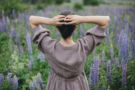 Stylish woman in rustic dress relaxing among lupine meadow, atmospheric image. Cottagecore aesthetics. Young female in linen dress gathering flowers in summer countryside, rural slow lifeの写真素材