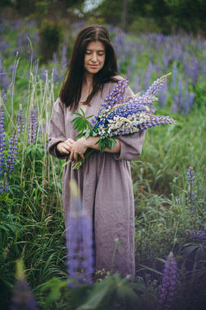 Cottagecore aesthetics. Stylish woman in rustic dress gathering lupine bouquet in meadow. Young female in linen dress with wildflowers in atmospheric summer countryside, rural slow lifeの写真素材