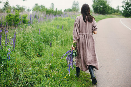 Stylish woman in rustic dress walking with lupine bouquet in summer countryside. Cottagecore aesthetics. Young female in linen dress holding wildflowers after rain on rural road, slow lifeの写真素材