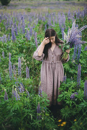 Stylish woman in rustic dress holding lupine bouquet in meadow. Cottagecore aesthetics. Young female in linen dress gathering wildflowers in atmospheric summer countryside, rural slow lifeの写真素材
