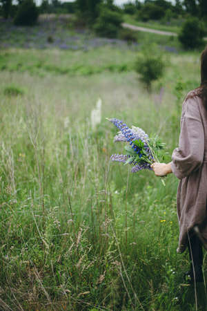 Close up of woman holding lupine bouquet in meadow. Cottagecore aesthetics. Young female in linen dress walking with wildflowers in atmospheric summer countryside, rural slow lifeの写真素材