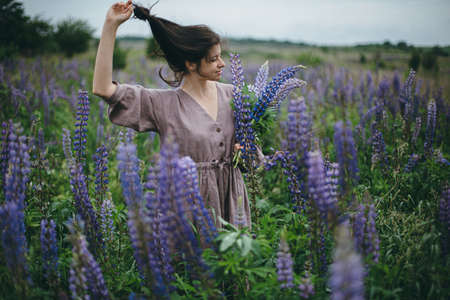 Stylish carefree woman in rustic dress holding lupine bouquet in meadow. Cottagecore aesthetics. Young female in linen dress gathering wildflowers in atmospheric summer countryside, slow lifeの写真素材