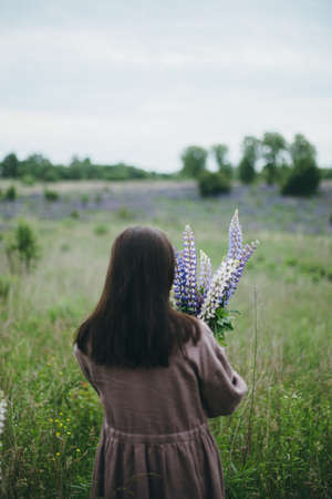 Stylish woman in rustic dress holding lupine bouquet in meadow. Cottagecore aesthetics. Young female...