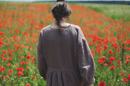 Stylish woman in rustic dress walking in red poppy field. Cottagecore aesthetics. Young female in linen dress relaxing among wildflowers meadow in sunny summer countryside, rural slow lifeの写真素材