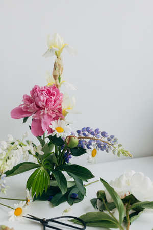 Modern summer flowers composition on rustic white table indoors. Creative floral image. Stylish peony, lupin,iris and daisy arrangement on kenzan with scissors on rustic wood.の写真素材