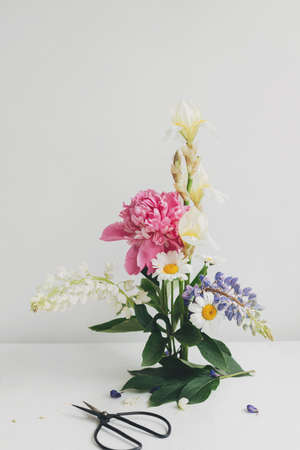 Modern summer flowers composition on rustic white table indoors. Creative floral image. Stylish peony, lupin,iris and daisy arrangement on kenzan with scissors on rustic wood.の写真素材