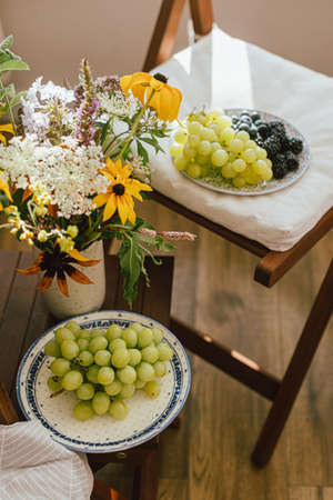 Fresh blueberries, blackberries and grapes in sunlight and wildflowers bouquet in rustic room. Summer berries in light, healthy food aesthetics. Summertime in countrysideの写真素材