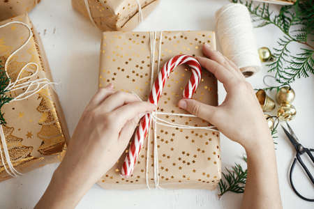 Hands wrapping stylish christmas gift flat lay. Person preparing modern christmas gift box with candy cane, festive paper, golden ornaments composition on white wood. Happy Holidays!の写真素材