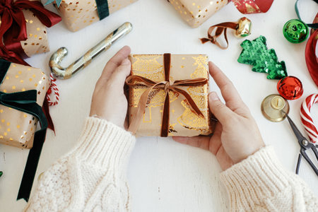 Hands wrapping stylish christmas gift. Person preparing modern gift box with velvet ribbon, golden wrapping paper, ornaments on white background. Merry Christmas! Atmospheric winter timeの写真素材