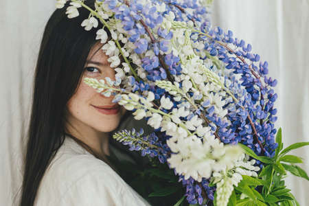Portrait of sensual woman holding lupine bouquet in rustic room. Gathering and arranging summer wildflowers at home in countryside. Stylish female in linen dress posing with lupine flowersの写真素材