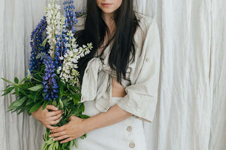 Stylish woman holding lupine bouquet in rustic room, close up. Young female in linen dress posing with lupine flowers. Gathering and arranging summer wildflowers at home in countrysideの写真素材