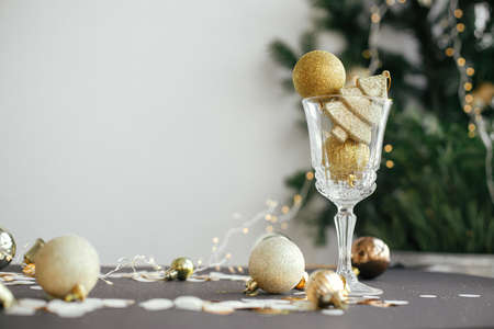 Modern christmas composition, golden glitter baubles in champagne glass and shiny decorations on dark table. Merry Christmas and Happy New Year! Moody holiday banner, creative still lifeの写真素材