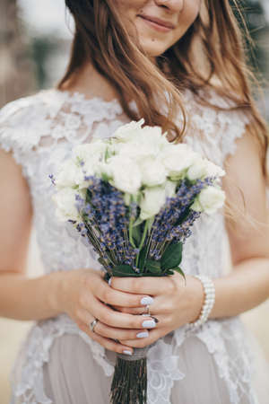 Portrait of sensual bride with wedding bouquet of roses and lavender in stylish gown. Provence wedding. Beautiful bride in stylish vintage dress posing in sunny street of european cityの写真素材