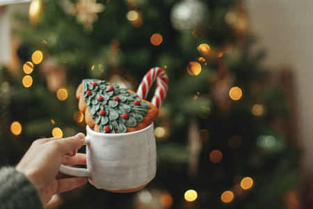Hand holding cup with cookie and candy cane on background of christmas tree with golden lights. Cozy winter at home. Woman holding tasty drink and sweets at illuminated christmas treeの写真素材