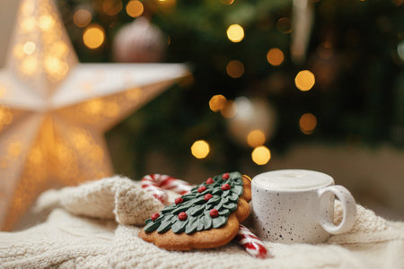 Warm cup of coffee, christmas tree gingerbread cookie and candy cane on knitted sweater on background of christmas tree with lights. Atmospheric winter hygge. Christmas bannerの写真素材