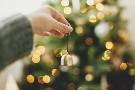 Hand holding golden bell on background of illuminated christmas tree with golden lights. Woman decorating christmas tree and holding glitter ornament in hand in festive room. Atmospheric timeの写真素材