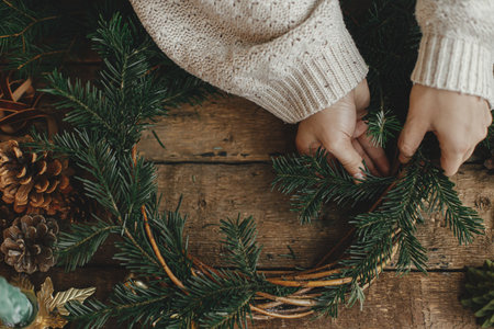 Making Christmas rustic wreath. Woman hands holding fir branches and making wreath on rustic wooden table with ribbon, golden bells, top view. Moody holiday image. Winter holiday workshopの写真素材