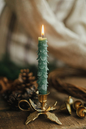 Stylish christmas candle as tree and golden bells on background of woman making christmas wreath on rustic wooden table. Making Christmas rustic wreath, moody image. Winter holiday workshopの写真素材