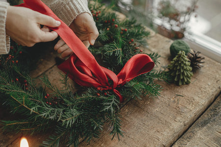 Making Christmas wreath. Woman hands decorating christmas wreath with red ribbon on rustic wooden table with pine cones and candles. Moody holiday image. Holiday workshop, winter decorの写真素材