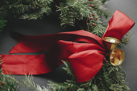 Stylish christmas wreath with red ribbon and golden bells on dark background close up. Modern winter holiday decor. Merry Christmas and Happy Holidays!の写真素材
