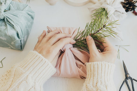 Zero waste Christmas concept. Hands decorating gift box wrapped in fabric with fir branch on white wooden table with eco friendly decor and candle. Furoshiki gift wrapping. Eco winter holidayの写真素材