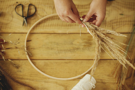 Hands arranging dried grass on hoop on wooden table with scissors, thread. Making stylish autumn...