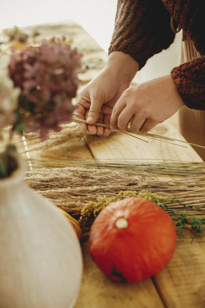 Making stylish autumn wreath on rustic table. Hands arranging dried grass on hoop on wooden table with scissors, thread, pumpkin. Fall decor and arrangement in farmhouse.の写真素材