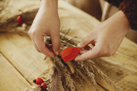 Making stylish autumn wreath on rustic table. Woman hands arranging berries, dry grass, herbs in wreath on wooden table. Fall decor and arrangement in farmhouseの写真素材
