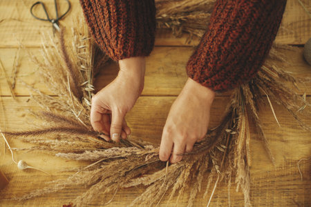 Woman hands arranging dried grass in wreath on wooden table, top view. Making stylish autumn wreath on rustic table. Fall decor and arrangement in farmhouseの写真素材