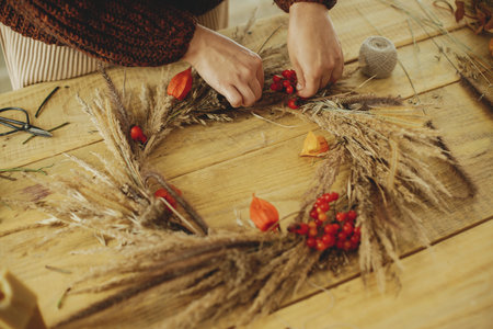 Making stylish autumn wreath on rustic table. Woman hands arranging berries, dry grass, herbs in...