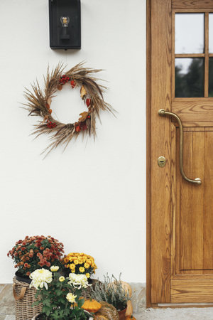 Stylish autumn wreath hanging at modern lantern, pumpkins and flowers at wooden door outdoor. Fall decor and arrangement of farmhouse exterior. Rustic autumn decorationの写真素材