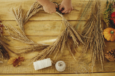 Making stylish autumn wreath on rustic table. Hands arranging dried grass on hoop on wooden table...