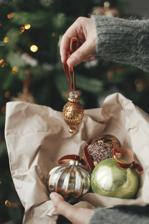 Hands decorating christmas tree with stylish bauble in atmospheric festive room. Merry Christmas! Winter holidays preparation. Woman in cozy sweater putting vintage toy on treeの写真素材