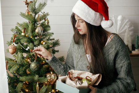 Stylish woman in cozy sweater and santa hat decorating christmas tree in atmospheric festive room. Merry Christmas! Winter holidays preparation. Decorating tree with vintage baublesの写真素材