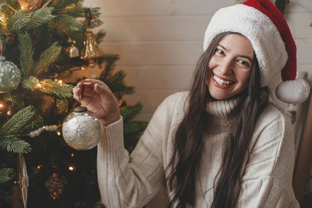 Merry Christmas! Happy woman in cozy sweater and santa hat decorating christmas tree with stylish baubles in atmospheric festive room. Winter holidays preparationの写真素材
