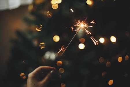 Happy New Year! Burning sparkler in female hand on background of christmas tree lights in dark room. Atmospheric celebration. Hand holding firework against stylish decorated tree with illuminationの写真素材