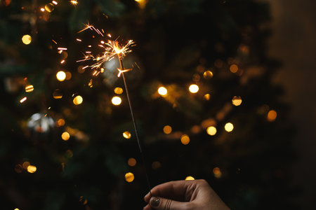 Burning sparkler in female hand on background of christmas tree lights in dark room. Happy New Year!...