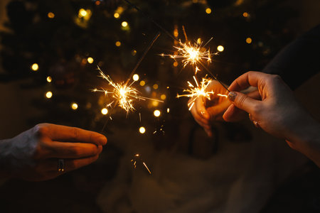 Friends celebrating with burning sparklers in hands against christmas lights in dark room. Happy New Year! Atmospheric holiday. Hands holding fireworks on background of stylish illuminated treeの写真素材