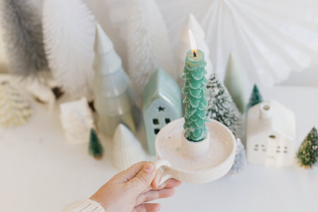 Merry Christmas! Hand holding stylish christmas tree candle on background of miniature winter village on white table. Modern little Christmas trees and houses. Holiday adventの写真素材