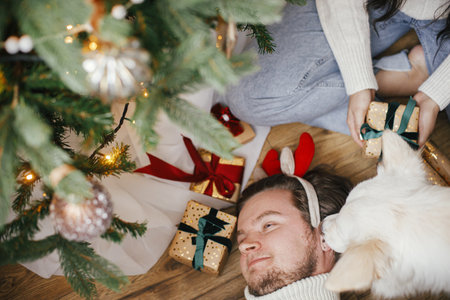 Cute happy dog playing with owner under stylish christmas tree with gifts, top view. Pet and winter holidays. Adorable danish spitz dog and man in reindeer antlers in festive room. Merry Christmas!の写真素材