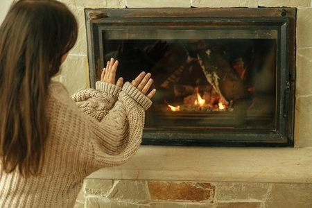 Heating house in winter with wood burning stove. Woman in cozy sweater warming up hands at fireplace in rustic room. Young stylish female sitting at fireplace in farmhouseの写真素材