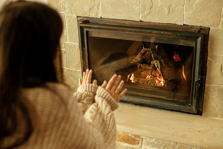 Heating house in winter with wood burning stove. Woman in cozy sweater warming up hands at fireplace in rustic room. Young stylish female sitting at fireplace in farmhouseの写真素材