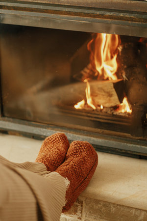 Heating house in winter with wood burning stove. Woman in cozy wool socks warming up feet at...
