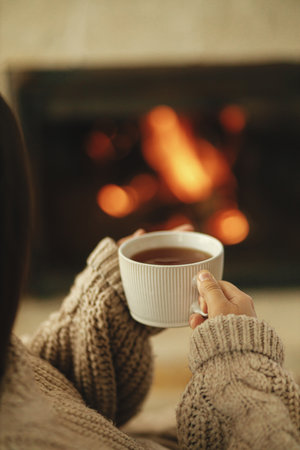 Hands in cozy sweater holding cup of warm tea on background of burning fireplace close up, autumn...